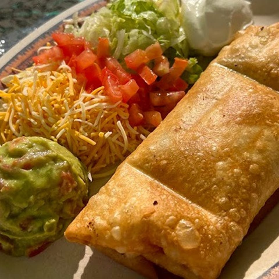 Crispy golden burrito served with guacamole, shredded cheese, diced tomatoes, lettuce, and sour cream on a patterned plate.